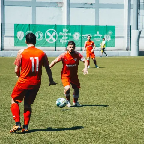 Spielszene: Ein Spieler in oranger Trikot mit Sponsor bautechnik führt den Ball, ein Gegenspieler mit Nummer 11 steht im Vordergrund, im Hintergrund die Bande.