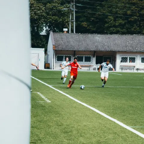 Spielszene: Ein Spieler in Rot dribbelt den Ball auf dem Rasen, während zwei Gegner in Weiß nachsetzen, im Hintergrund das Vereinsgebäude.