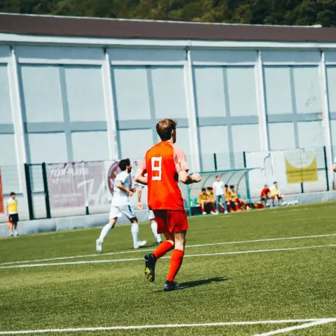 Spieler in orangem Trikot mit Rückennummer 9 läuft auf dem Kunstrasen, im Hintergrund Zuschauer auf der Bank und ein weißgekleideter Gegenspieler.