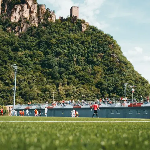 Fußballplatz mit Spielern in Weiß und Orange vor einer Tribüne, dahinter ein bewaldeter Felsen mit Burgturm, Spiel Terlan gegen Bruneck.