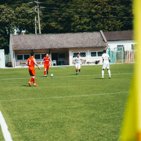 Spielszene: Ein Spieler in Orange führt den Ball am Mittelkreis vor, während Spieler in Weiß und Hellblau über das Feld laufen.