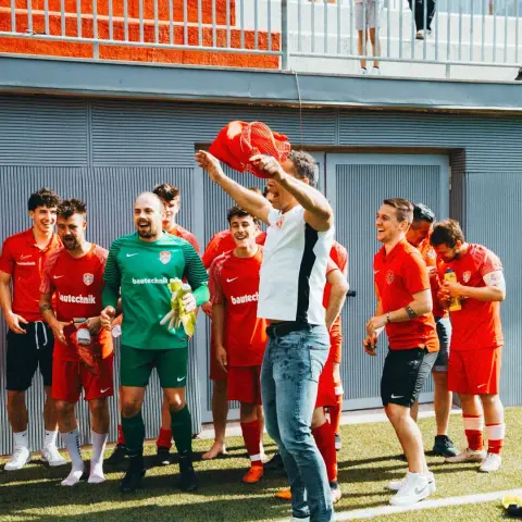 Ein Mann im weißen Shirt springt auf dem Rasen und hält ein rotes Trikot in die Höhe, während die lachende Mannschaft in Rot und Grün jubelt.